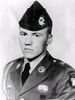 Head and shoulders of a young white man wearing a garrison cap with a large badge on the left side and a military jacket with a braided shoulder cord and several patches and pins on the lapels, shoulders, and chest.