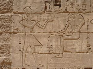 Relief of a man with a crown holding a tray of food in front of a seated man with the head of a ram