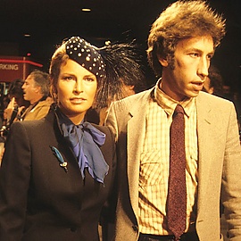 Welch in a blue scarf and high-collared gray jacket, with polka-dot feathered cap, in company of a man