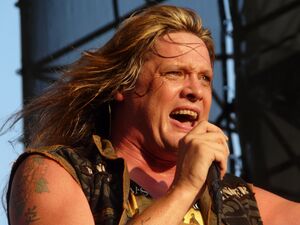 A middle-aged Caucasian man with long, blond hair and tattoos on his right shoulder wearing a sleeveless khaki shirt sings into a microphone.