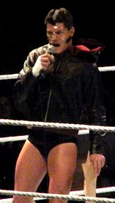 A dark-haired Caucasian male stands in a wrestling ring with grey ring ropes. He is wearing short black wrestling tights with a black, hooded jacket, and his face is covered with a clear plastic mask. He is holding a microphone to his mouth with his right hand, and in his left hand he is holding brown paper bags.