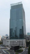 Ground-level view of a blue, glass high-rise; the tower sits behind a small, white, stone, window-dotted facade