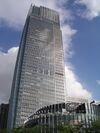 Ground-level view of a tall, mostly glass facade set in front of a blue, cloudy sky; a smaller, circular building is in the foreground